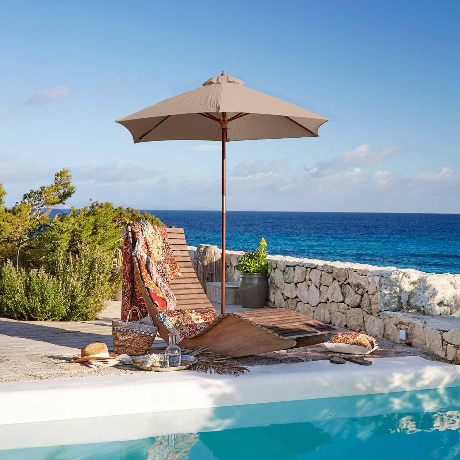 Chaise longue en bois avec parasol beige au bord de la piscine face à la mer, couverture colorée