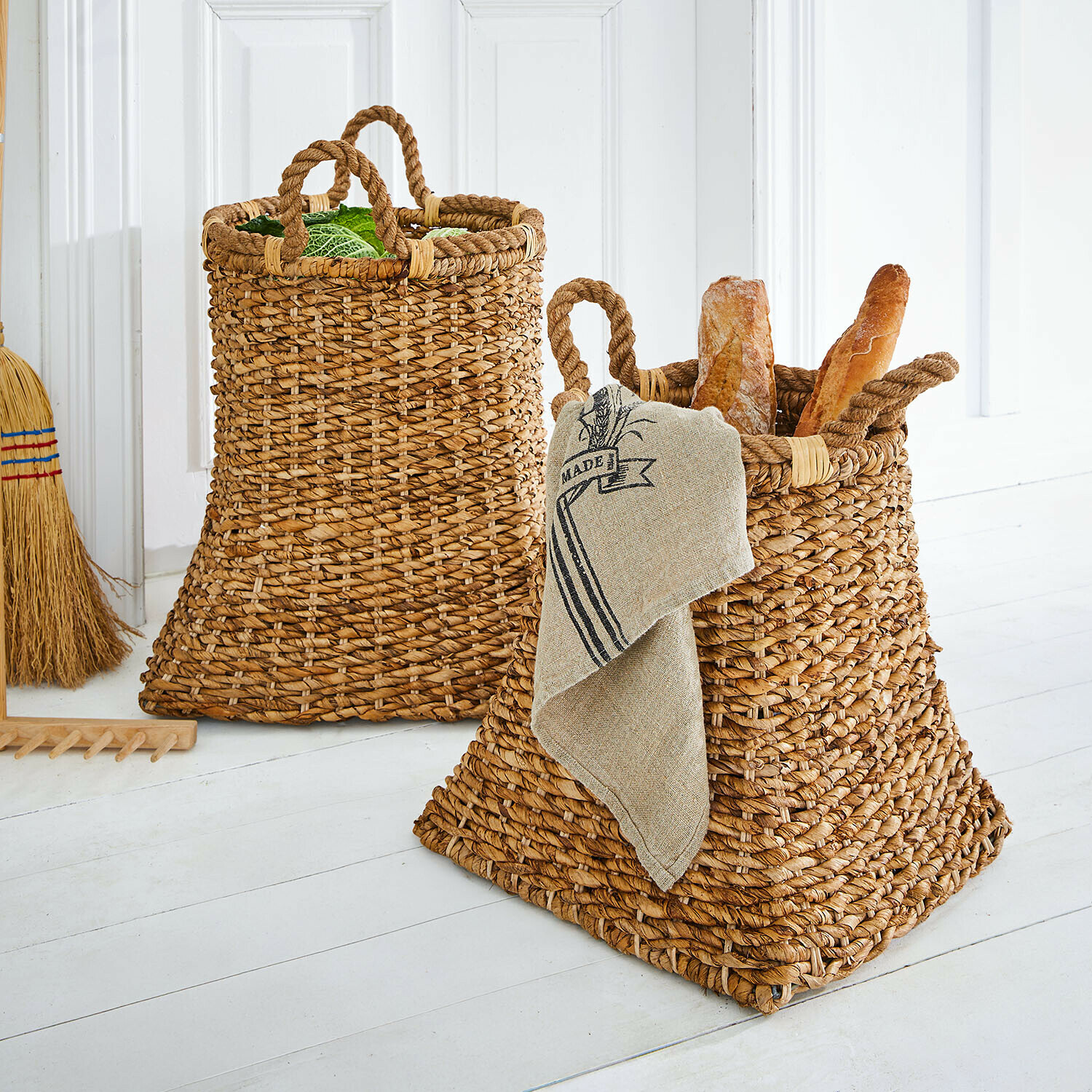 Lot de 2 paniers Saviani Deux paniers en osier naturel avec poignées, remplis de pain et de légumes verts dans un intérieur clair