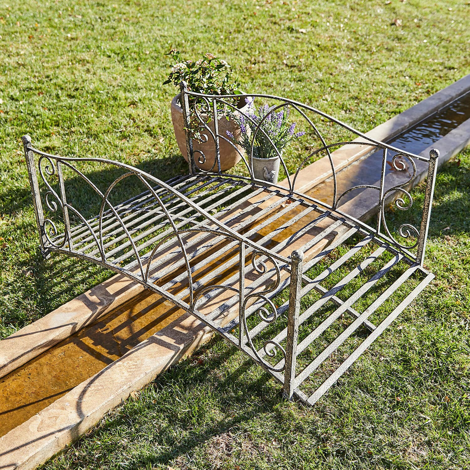 Pont décoratif Escàs Petit pont de jardin en métal vieilli argenté avec décorations, posé sur pelouse près d'un canal d'eau