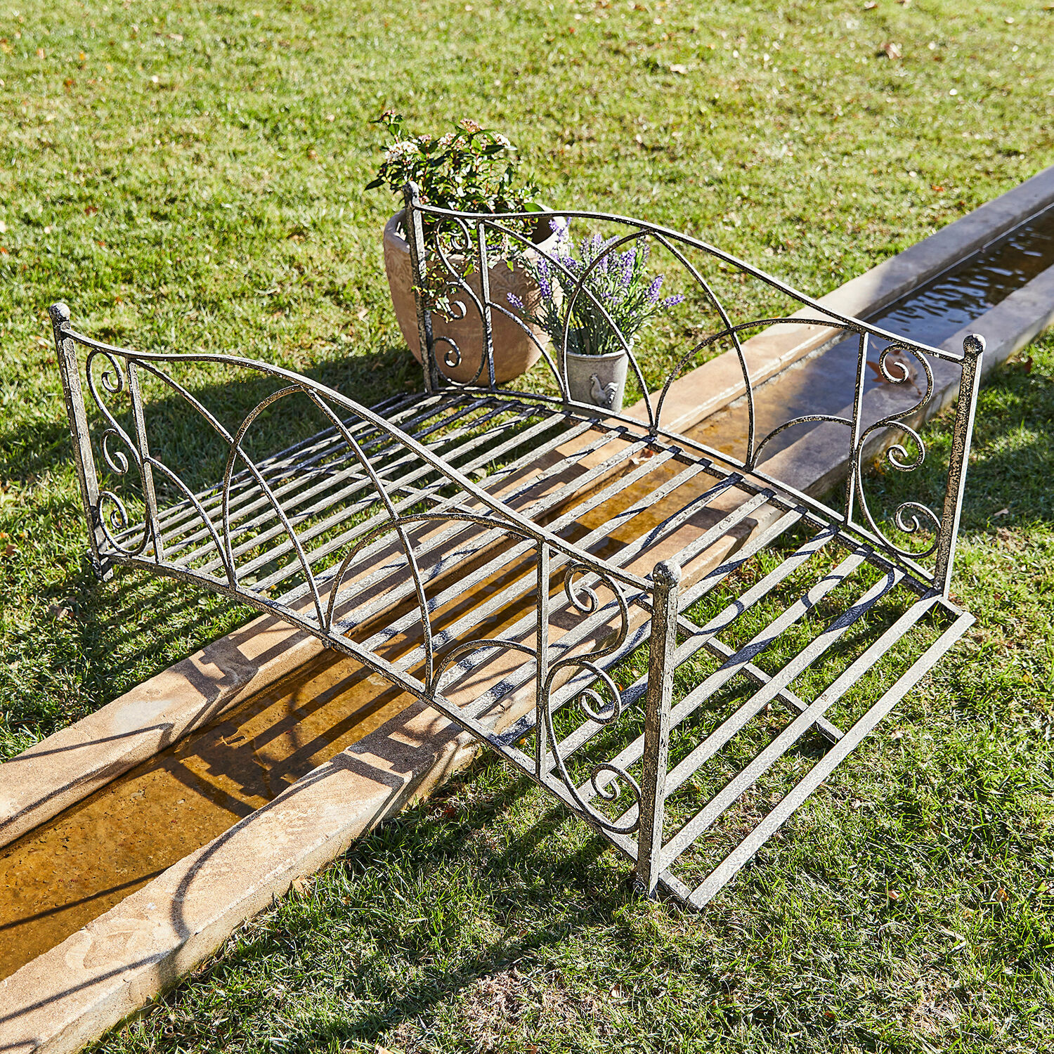 Pont décoratif Escàs Petit pont de jardin en métal vieilli argenté avec décorations, posé sur pelouse près d'un canal d'eau