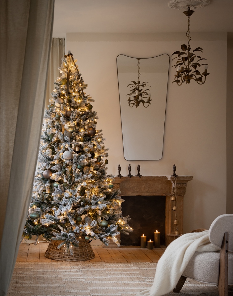 Sapin de Noël enneigé décoré de lumières et boules dans un salon chaleureux avec cheminée