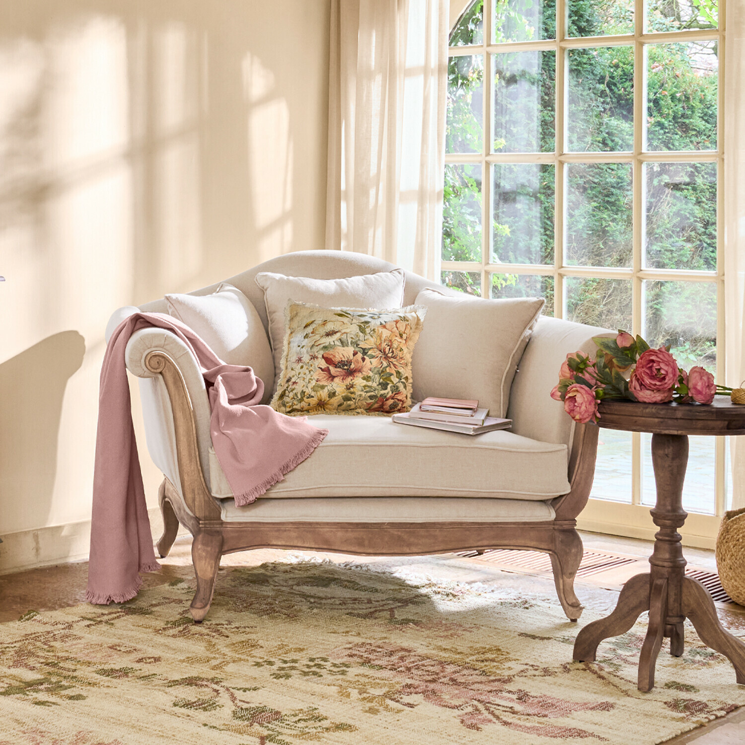 Fauteuil Sumerset Canapé beige en bois avec coussin floral et plaid rose dans un salon lumineux avec grande fenêtre