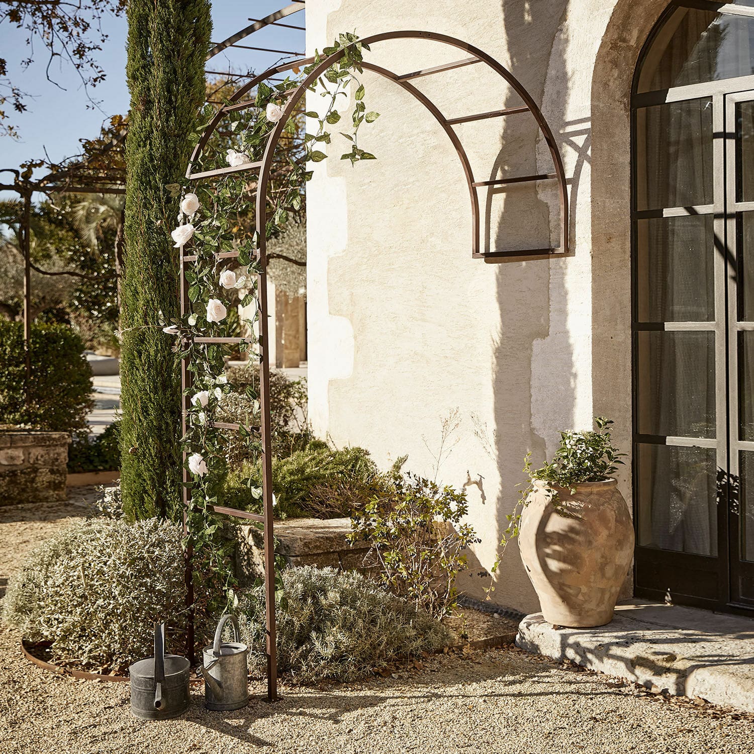 Arche à rosiers Auburn Pergola de jardin en métal brun avec arc décoré de roses blanches et arrosoirs sur gravier