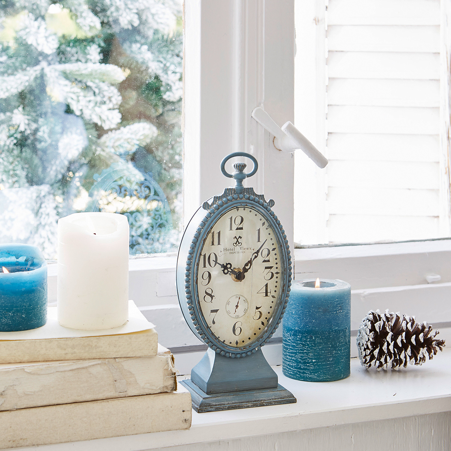 Horloge Michelet Horloge de table bleue en métal sur rebord de fenêtre blanc avec bougies bleues et blanches et pomme de pin hivernale