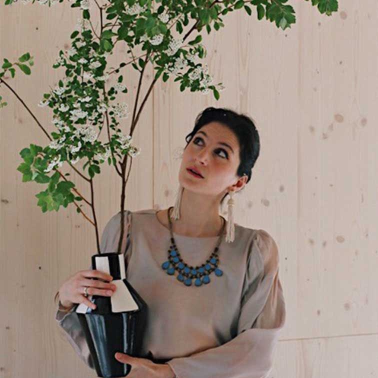 Femme tenant un vase en céramique noir et blanc avec des branches vertes devant un mur en bois clair