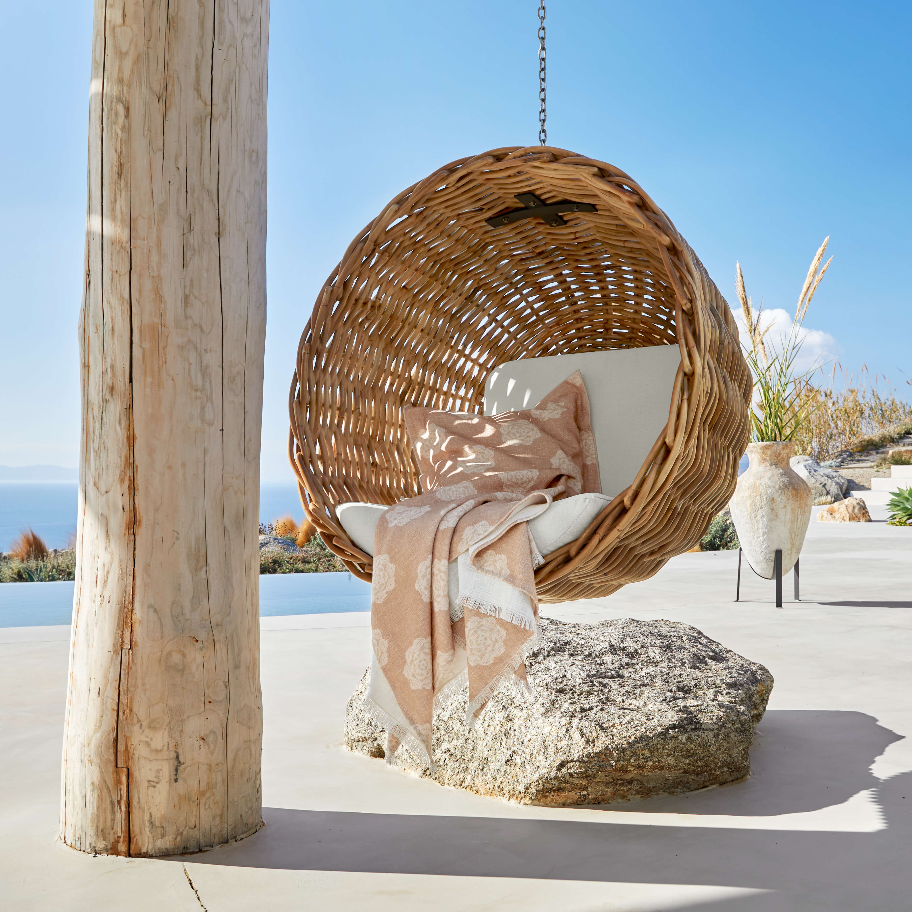 Fauteuil suspendu en rotin naturel avec coussin clair et plaid beige sur terrasse avec vue mer