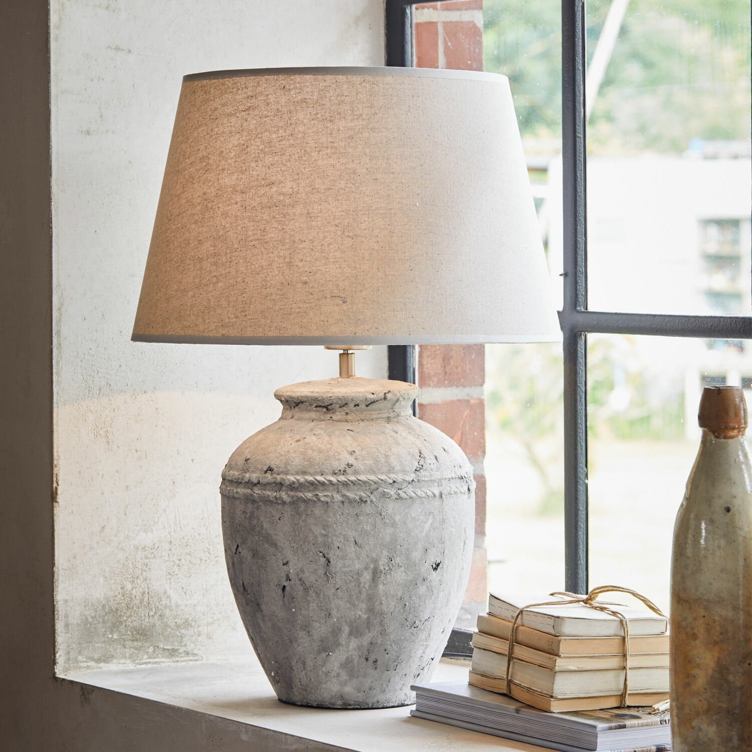 Lampe à poser Mosnay Lampe de table en céramique grise avec abat-jour beige posée sur un rebord de fenêtre avec livres