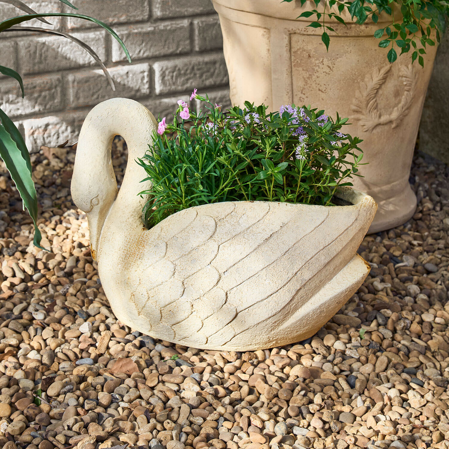 Cache-pot Belmarie Jardinière en forme de cygne en pierre claire avec plantes vertes et fleurs violettes sur gravier