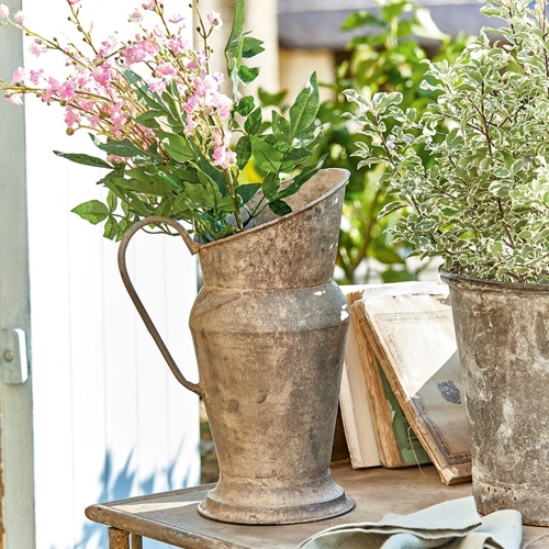 Pichet décoratif en métal vieilli avec fleurs vertes et roses sur une table de jardin ensoleillée