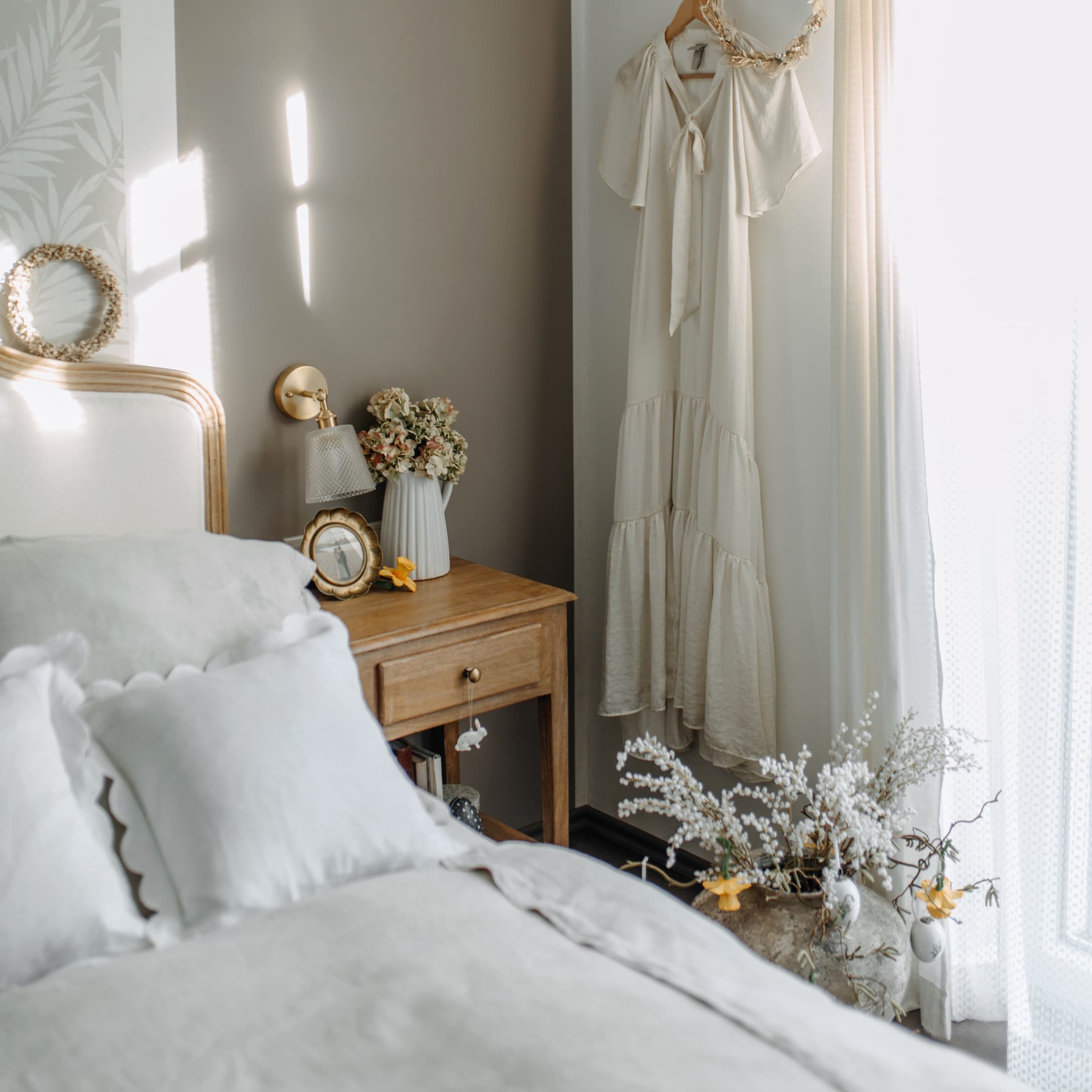 Chambre chaleureuse avec table de chevet en bois, literie crème et robe claire accrochée au mur
