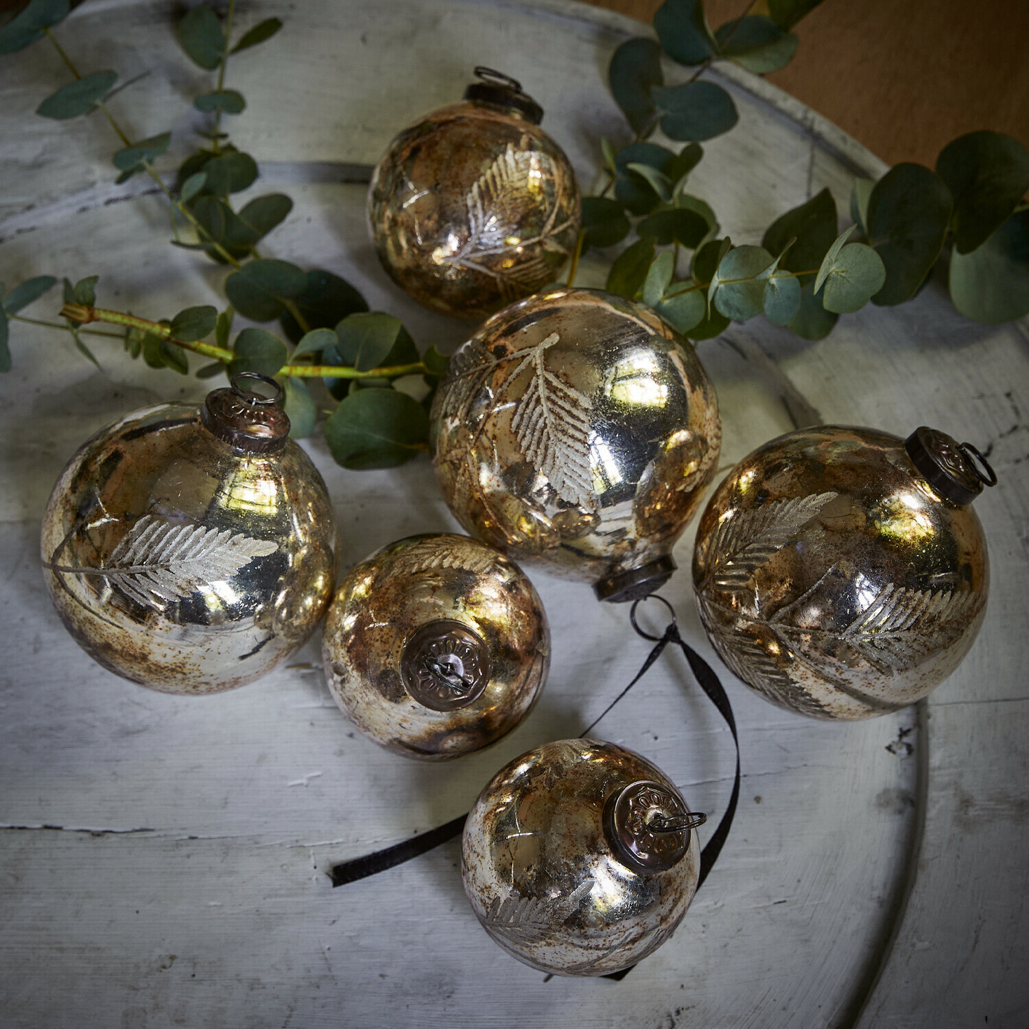 Lot de 6 décorations de Noël Tristan Ensemble de six boules de Noël en verre doré avec motif de feuilles sur un plateau blanc avec des branches vertes