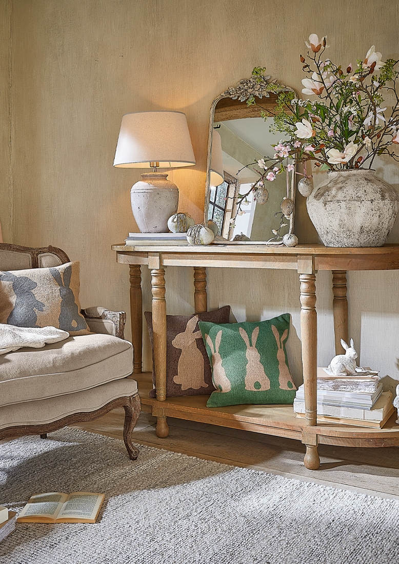 Console en bois avec lampe, miroir et décorations rustiques, coussins avec motifs lapins dans un salon cosy.