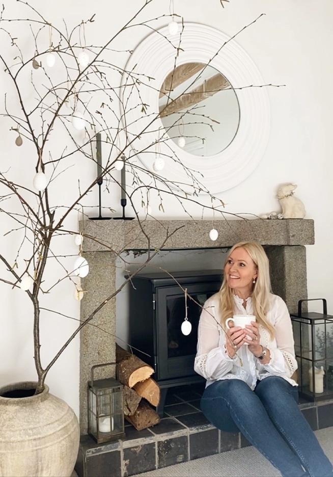 Femme assise près d'une cheminée en pierre avec branches décoratives et œufs blancs dans un intérieur chaleureux