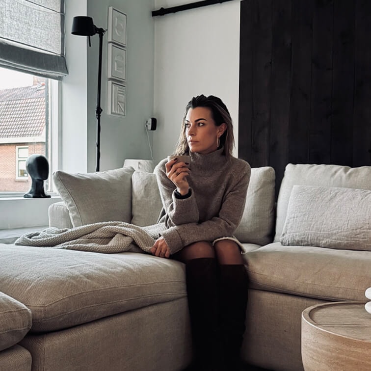 Femme en pull gris assise sur un canapé beige dans un salon moderne avec une grande fenêtre