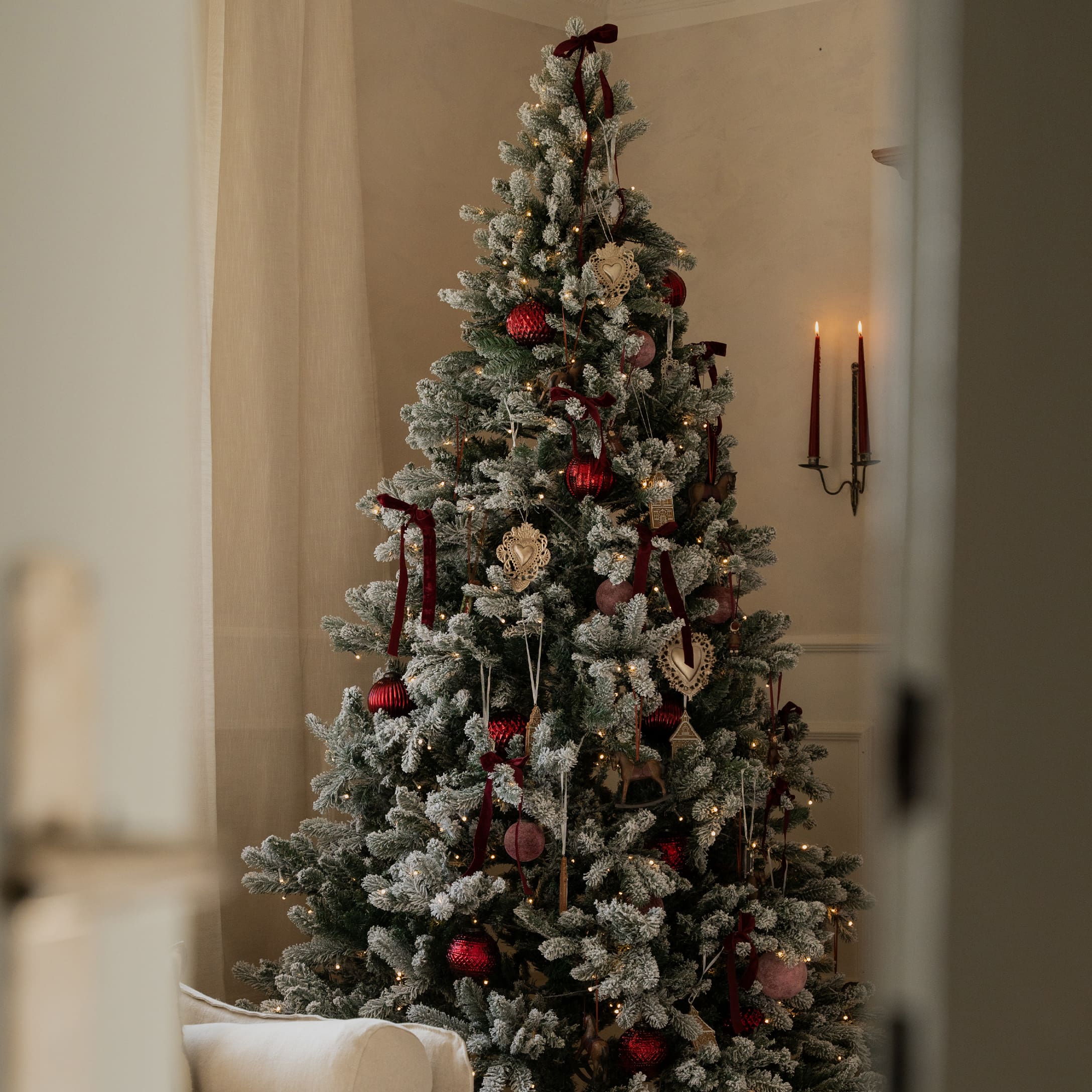 Sapin de Noël enneigé décoré de boules rouges et beiges avec lumières chaudes dans un intérieur cosy