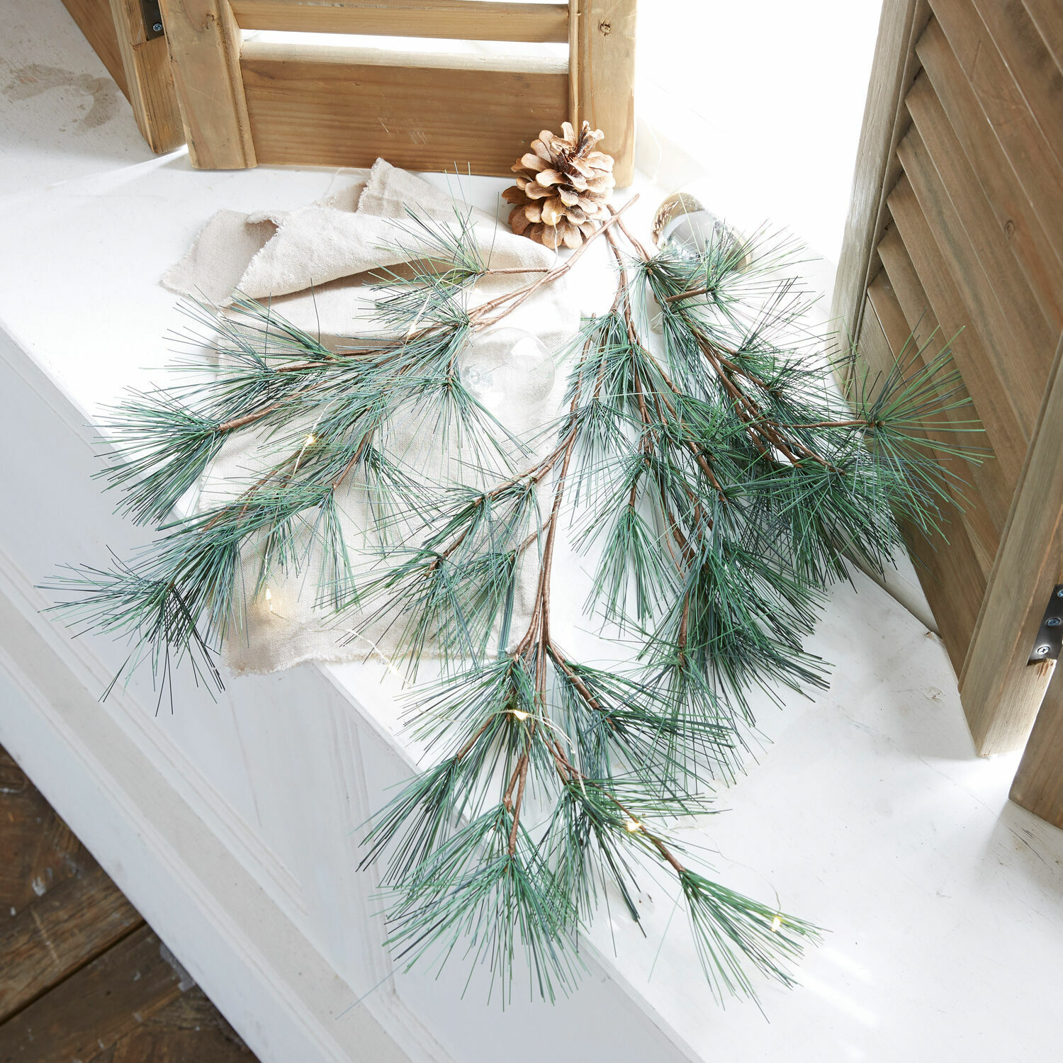 Lot de 4 branches Bohicon Branches de pin vertes avec aiguilles et guirlande LED posées sur un rebord blanc avec pomme de pin