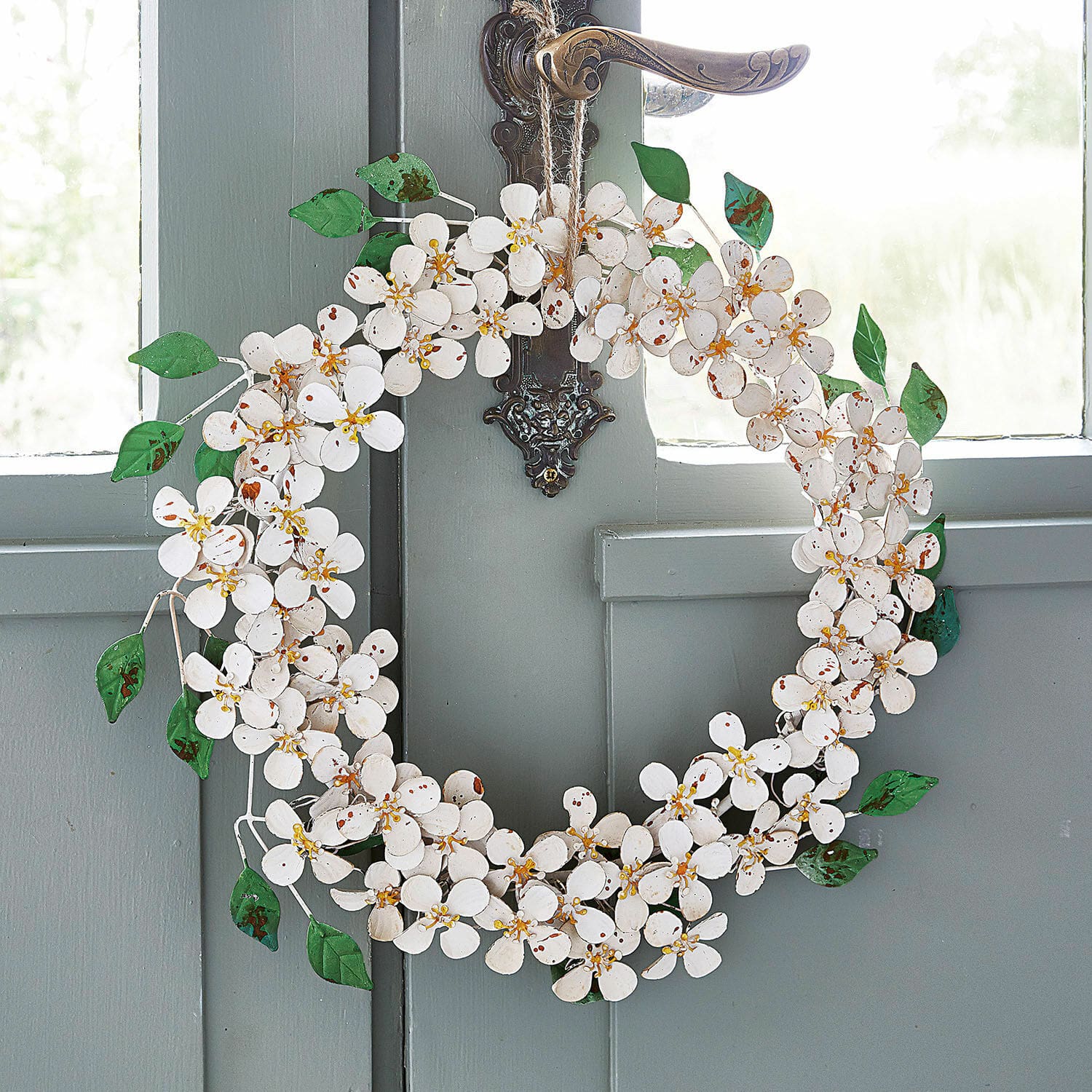 Couronne Loanne Couronne décorative en métal blanc avec fleurs et feuilles vertes suspendue à une porte grise