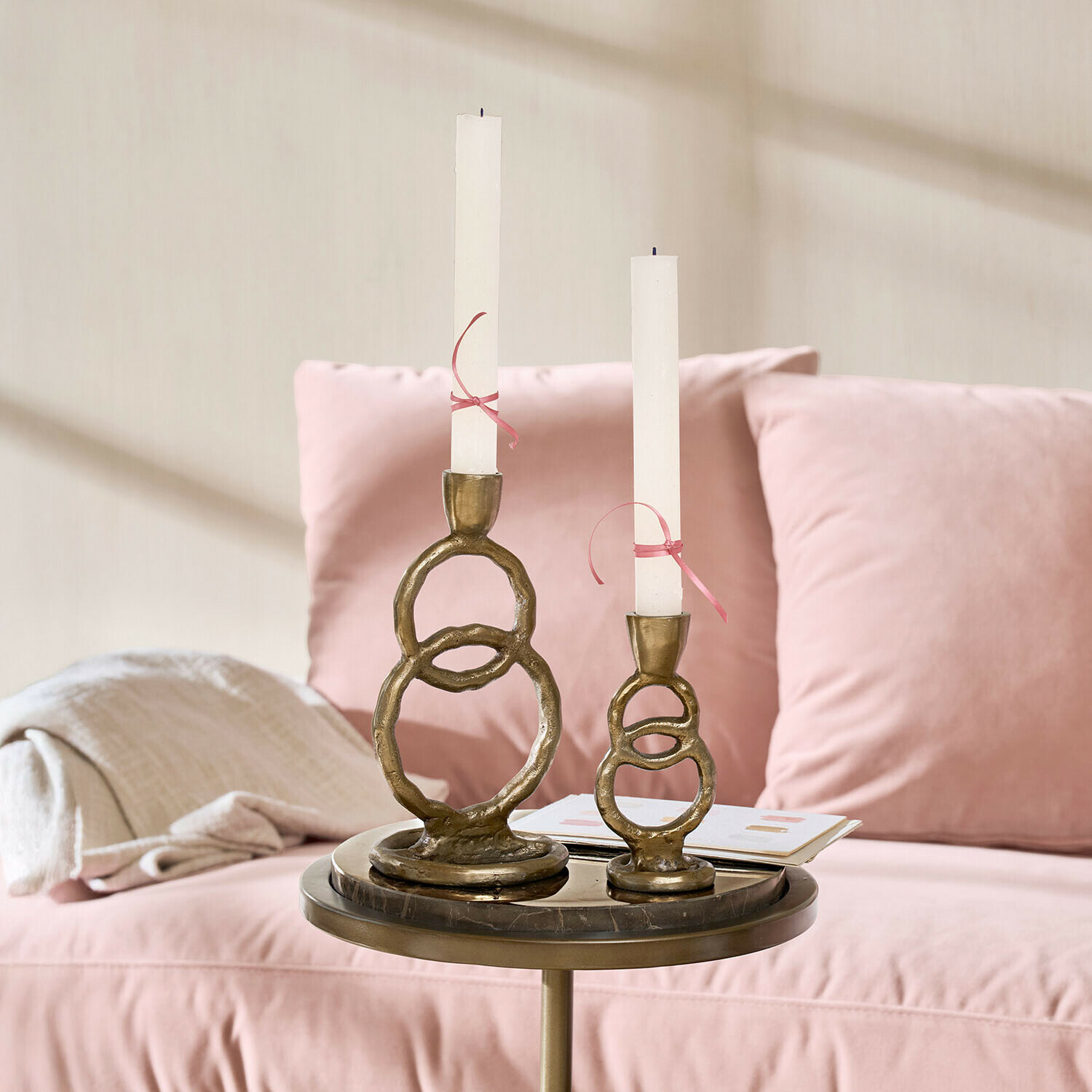 Lot de 2 bougeoirs Corvane Ensemble de deux bougeoirs en laiton avec bougies blanches sur une table près d'un canapé rose
