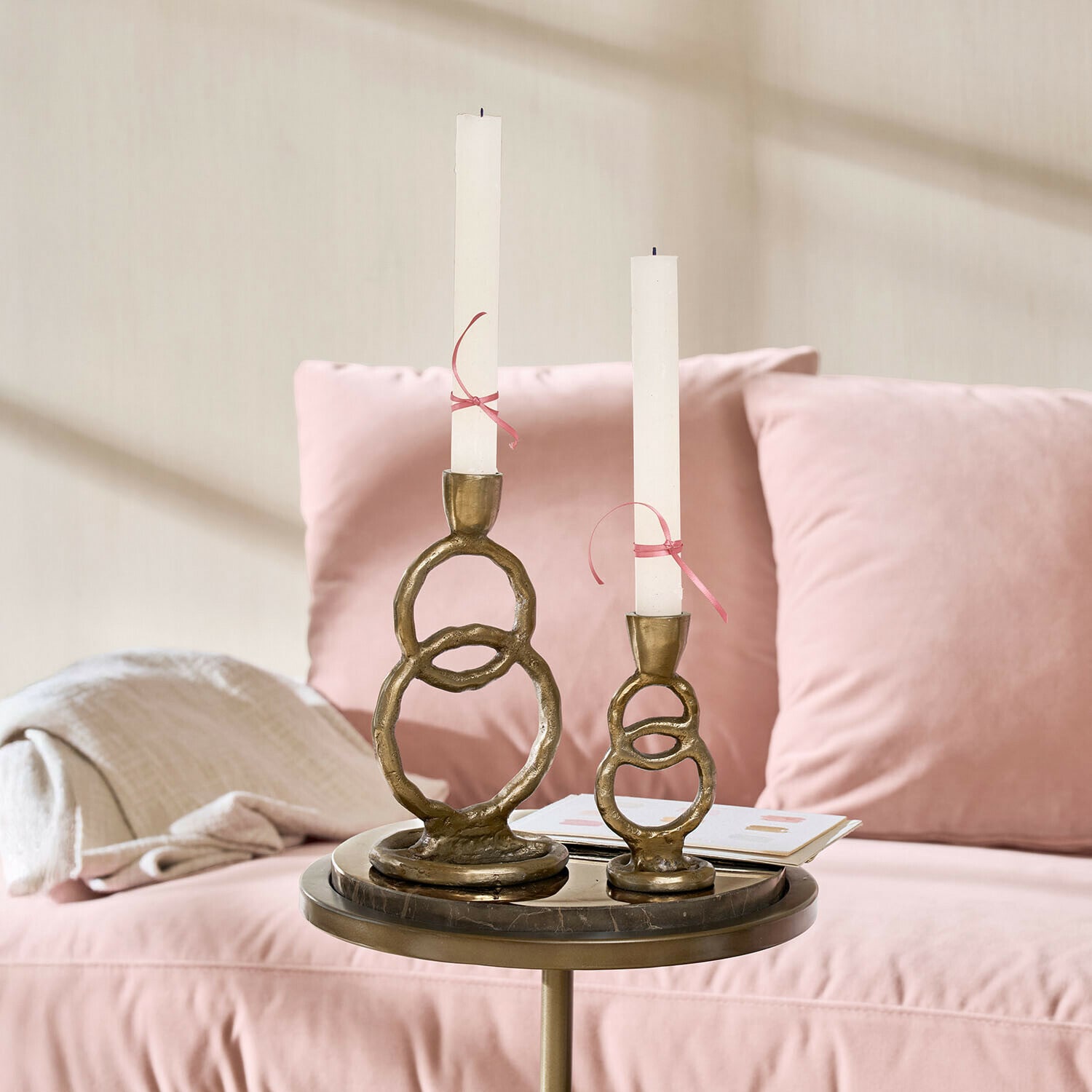 Lot de 2 bougeoirs Corvane Ensemble de deux bougeoirs en laiton avec bougies blanches sur une table près d'un canapé rose
