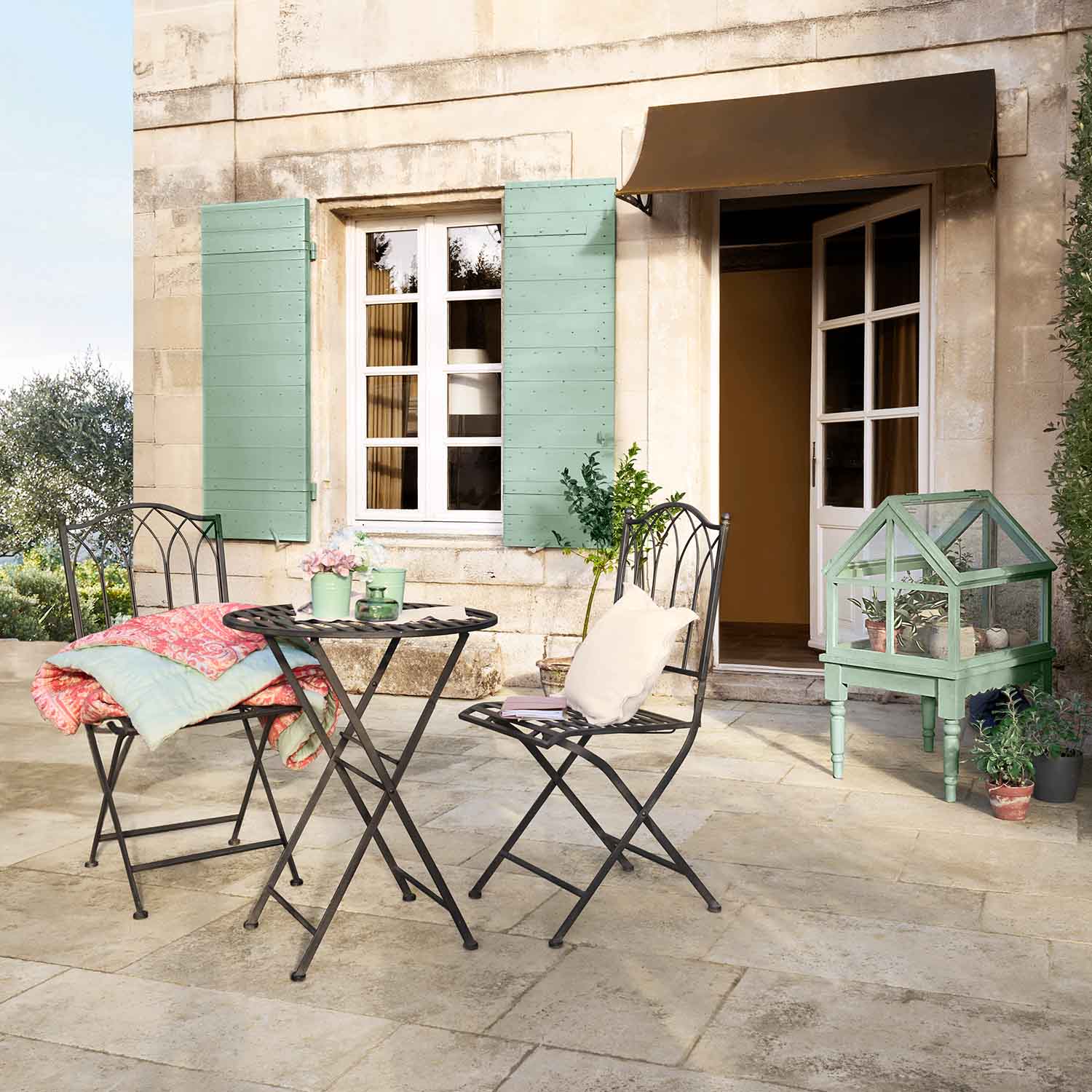 Ensemble de jardin en métal avec table et chaises sur terrasse, mini serre verte et coussins pastel