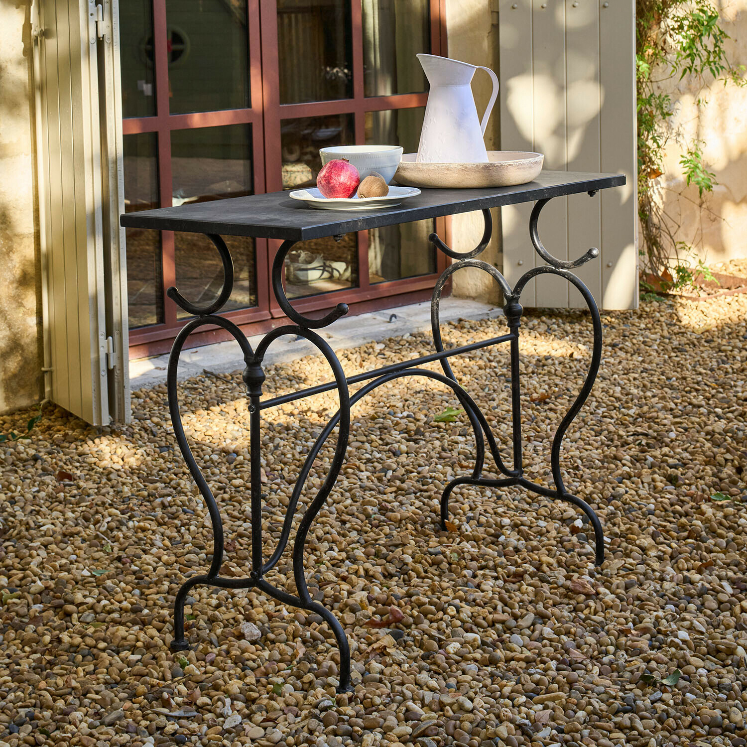 Table Cauvrel Table de jardin en métal noir avec plateau rectangulaire, posée sur un sol en gravier avec décorations