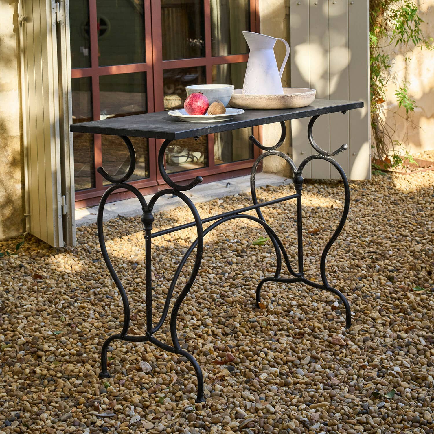 Table Cauvrel Table de jardin en métal noir avec plateau rectangulaire, posée sur un sol en gravier avec décorations