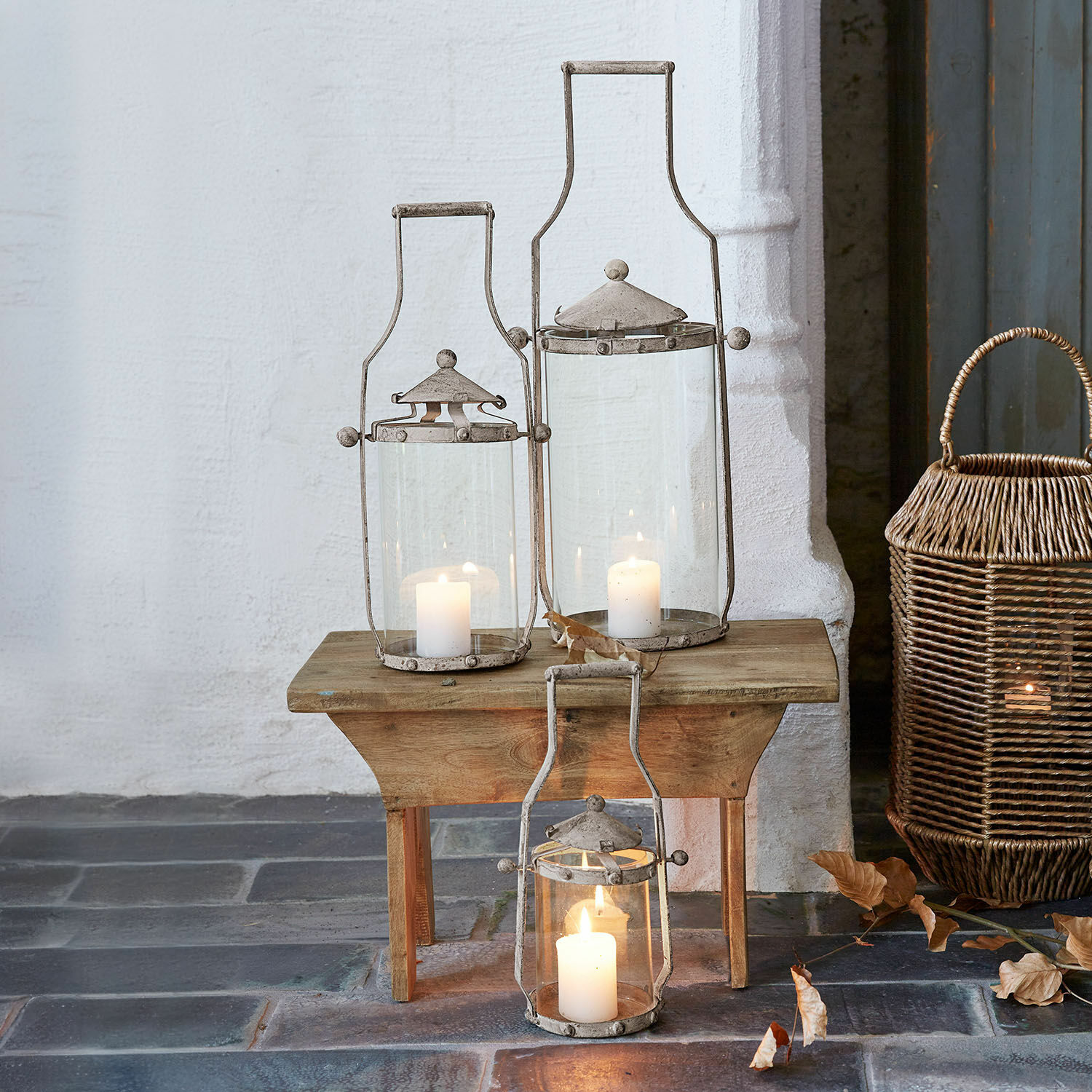 Lot de 3 lanternes Nery Ensemble de trois lanternes en métal gris avec bougies blanches sur une table en bois et sol en pierre