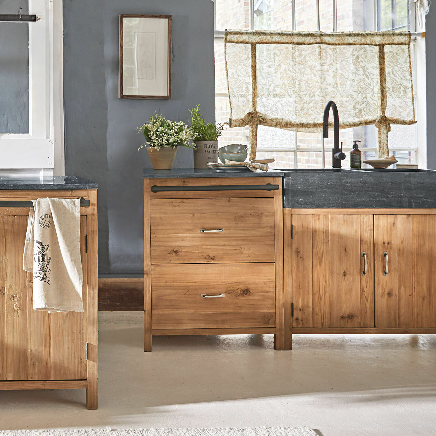 Meuble bas Sunnybrook Cuisine avec meubles en bois clair et plan de travail noir, décorations végétales et rideaux à motifs floraux
