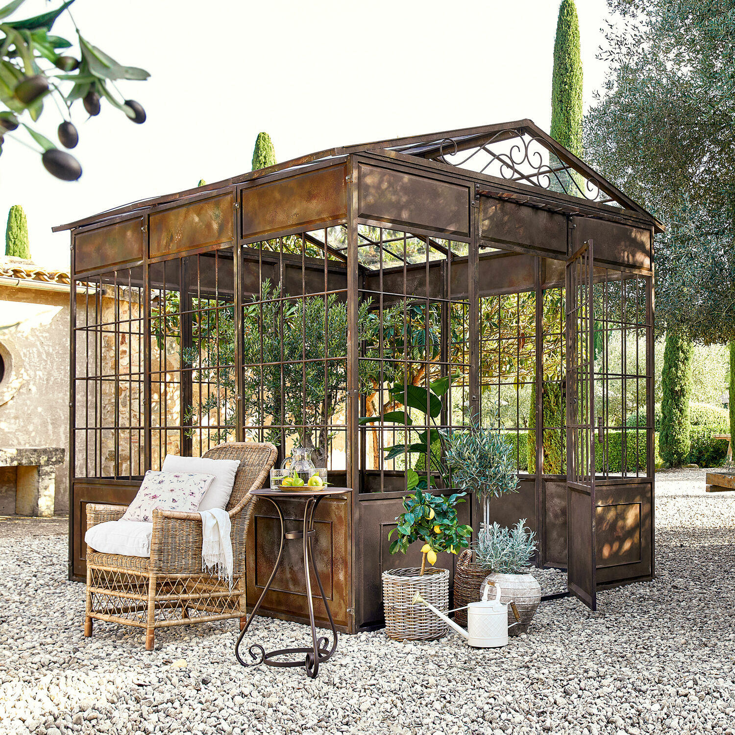 Gloriette Essone Orangerie en métal style rustique sur sol en gravier avec fauteuil en osier et plantes décoratives