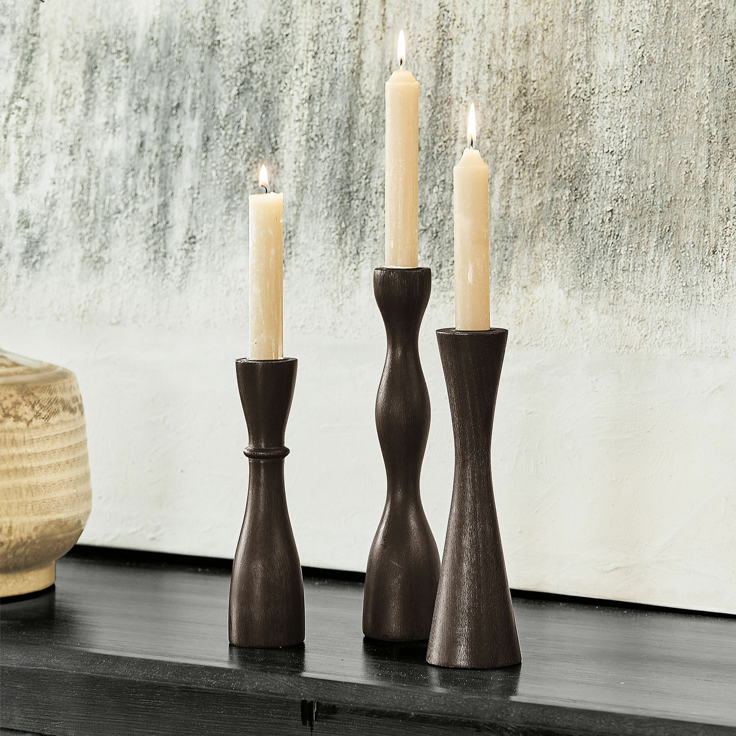 Lot de 3 bougeoirs Méléandre Ensemble de trois bougeoirs en bois foncé avec bougies crème sur surface noire