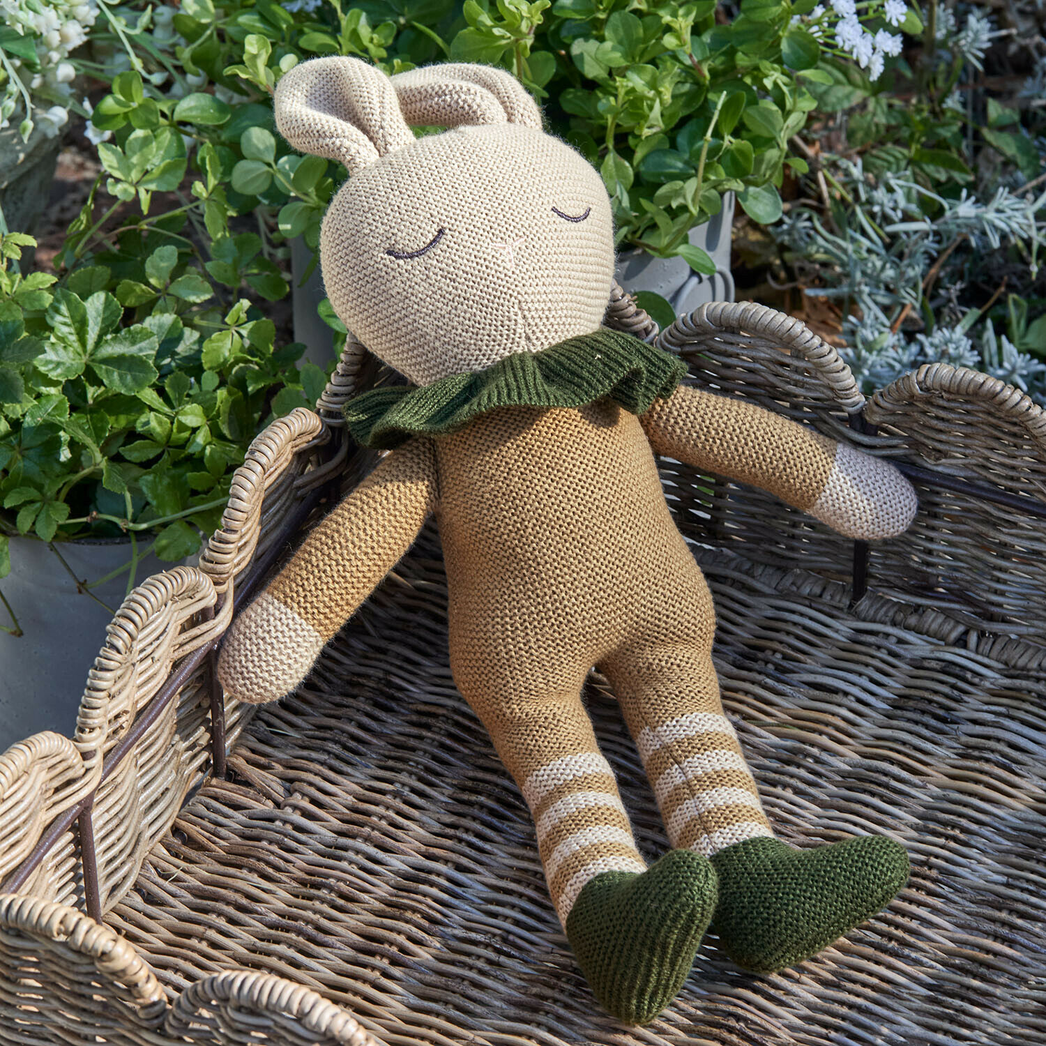 Peluche Cuddlecloud Jouet lapin tricoté beige avec chaussures et col verts posé sur un panier en osier entouré de plantes
