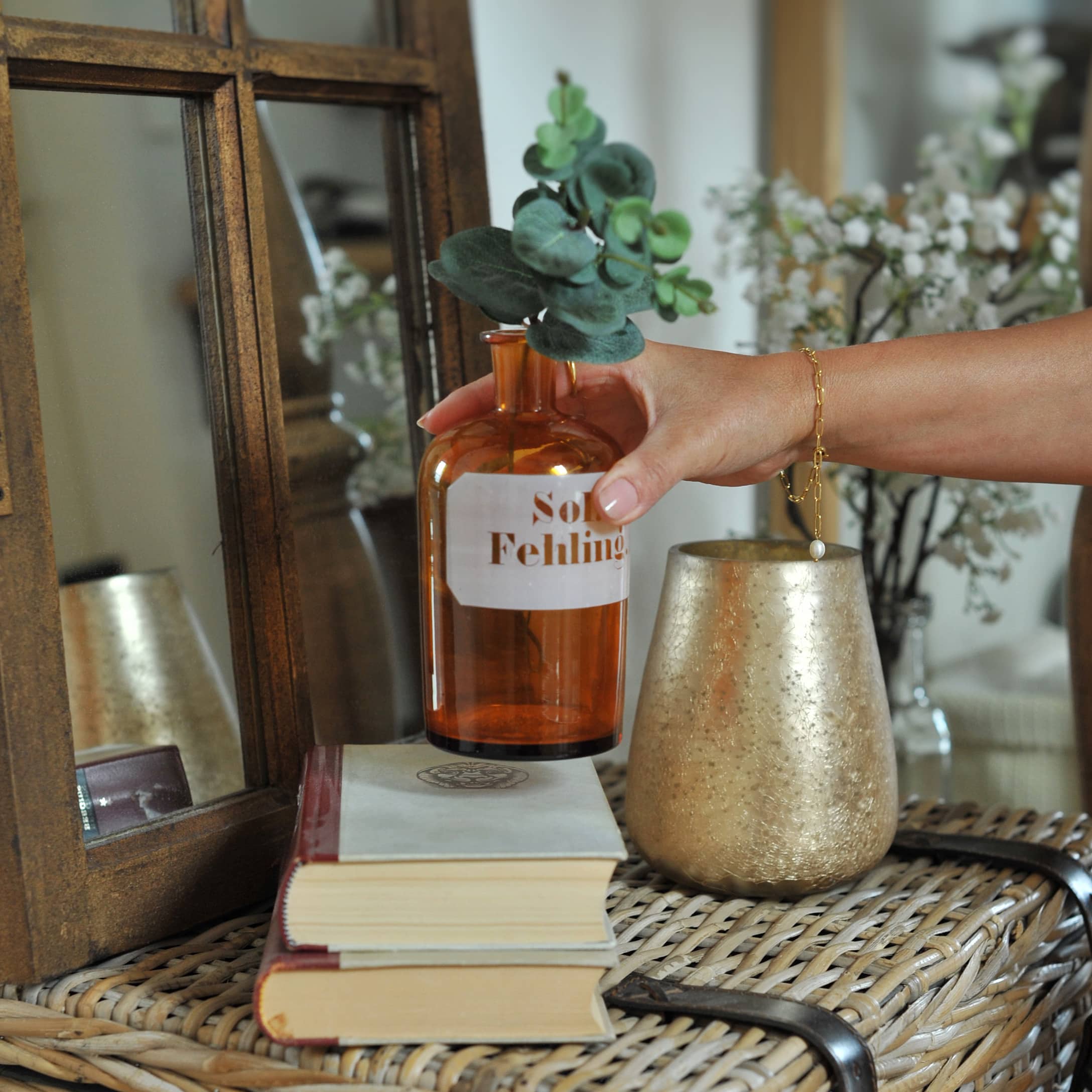 Main tenant un flacon en verre ambré avec feuilles vertes sur table en osier avec livres et vase doré