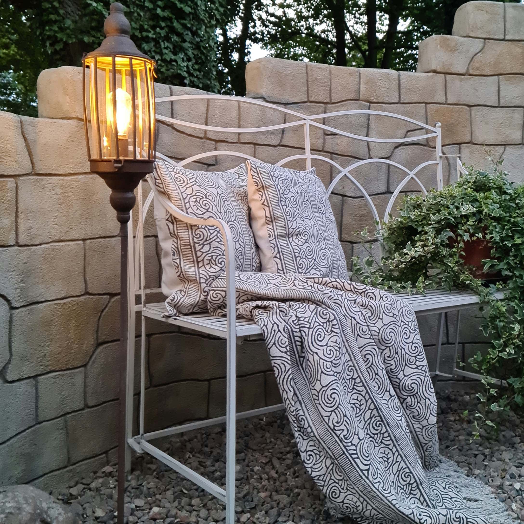 Banc de jardin en métal blanc avec coussins et plaid à motifs noirs et blancs, lampe sur pied allumée