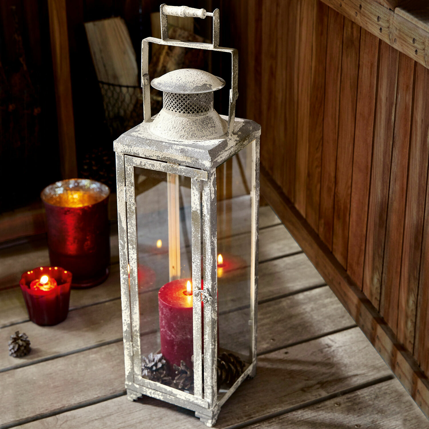Lanterne Isodore Lanterne en métal patinée blanc avec bougie rouge et pommes de pin sur plancher en bois