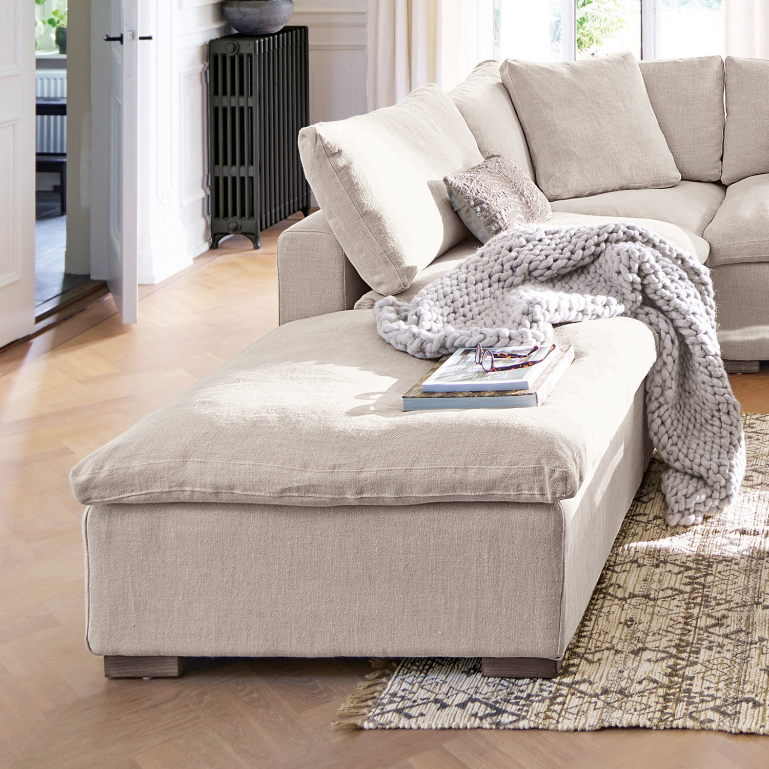 Pouf Bennington Canapé d'angle beige en tissu avec couverture douce et coussins dans un salon lumineux au sol en bois