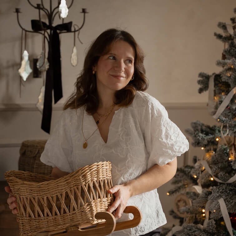 Femme en blouse blanche brodée tenant un panier en osier sur des patins en bois près d'un sapin de Noël décoré