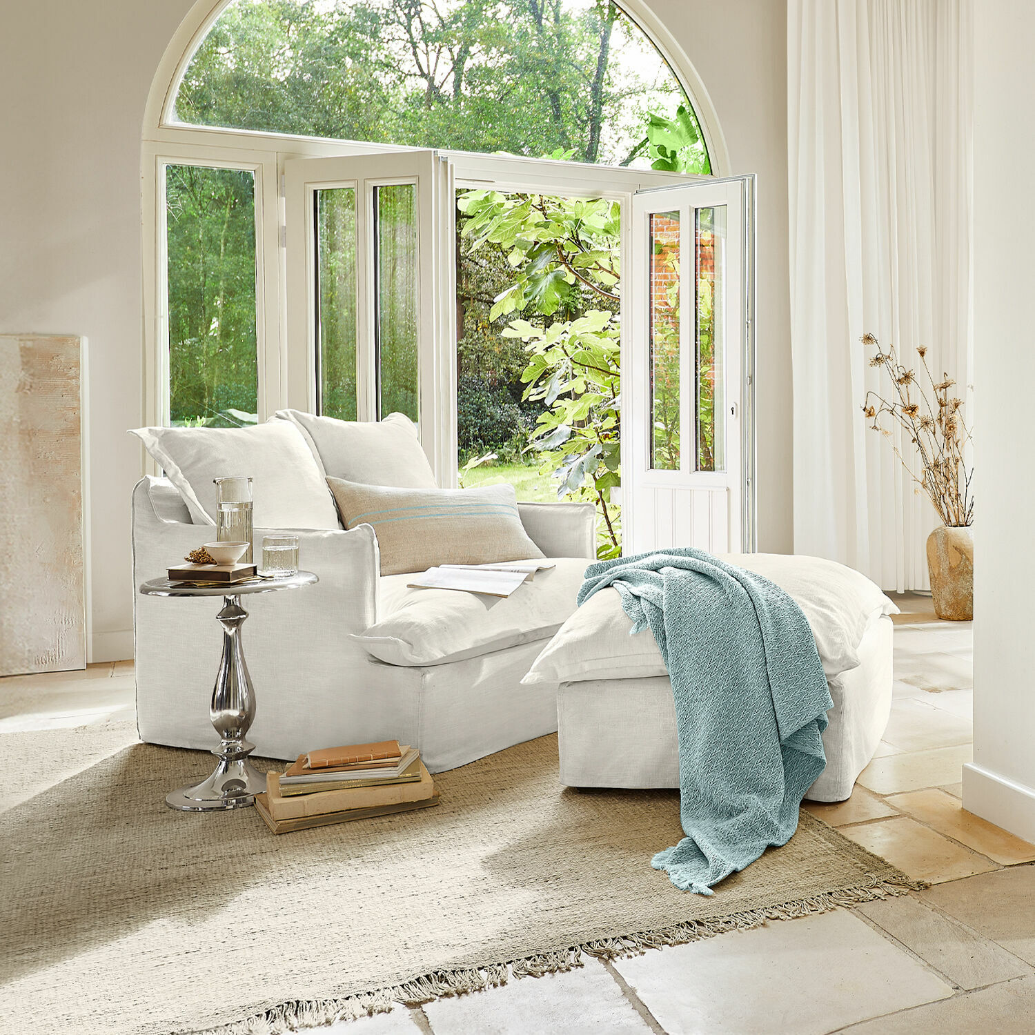 Fauteuil avec repose-pieds Avignon Fauteuil en coton blanc avec repose-pieds et couverture bleue dans un salon lumineux avec grande fenêtre