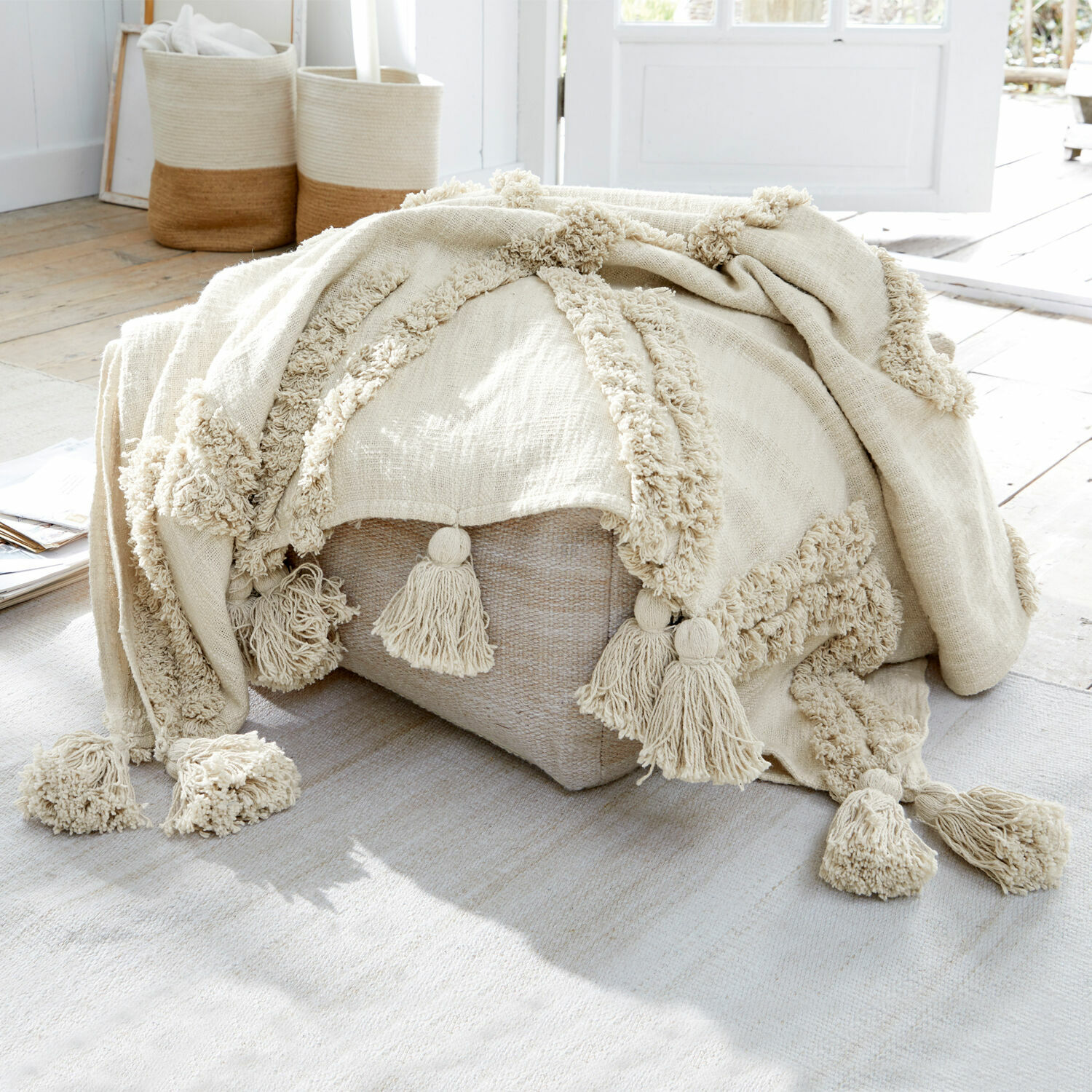 Plaid Linares Plaid beige en coton avec pompons sur un pouf clair dans un intérieur lumineux naturel