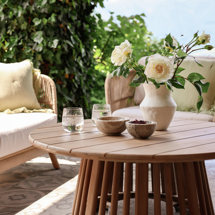 Table ronde en bois avec vase blanc en céramique et fleurs sur terrasse avec fauteuils en osier