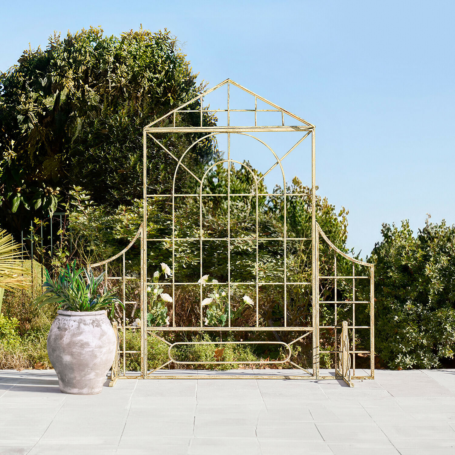 Treillis de jardin en métal doré en forme de maison sur terrasse avec pot et végétation en arrière-plan