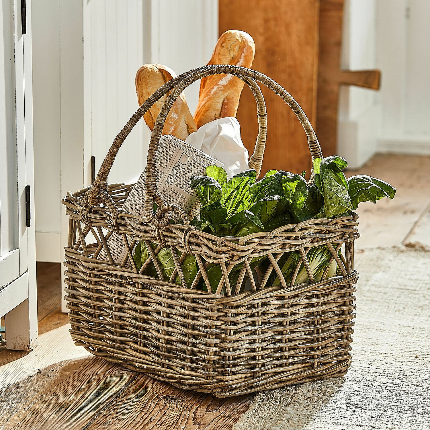 Corbeille Amixal Panier en osier avec poignées rempli de feuilles vertes et baguettes sur un sol en bois