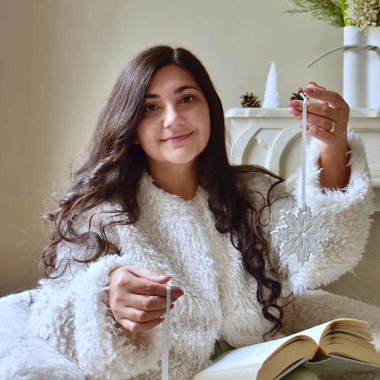 Femme en pull blanc duveteux tenant une décoration de Noël blanche en forme de flocon de neige avec un livre ouvert sur les genoux