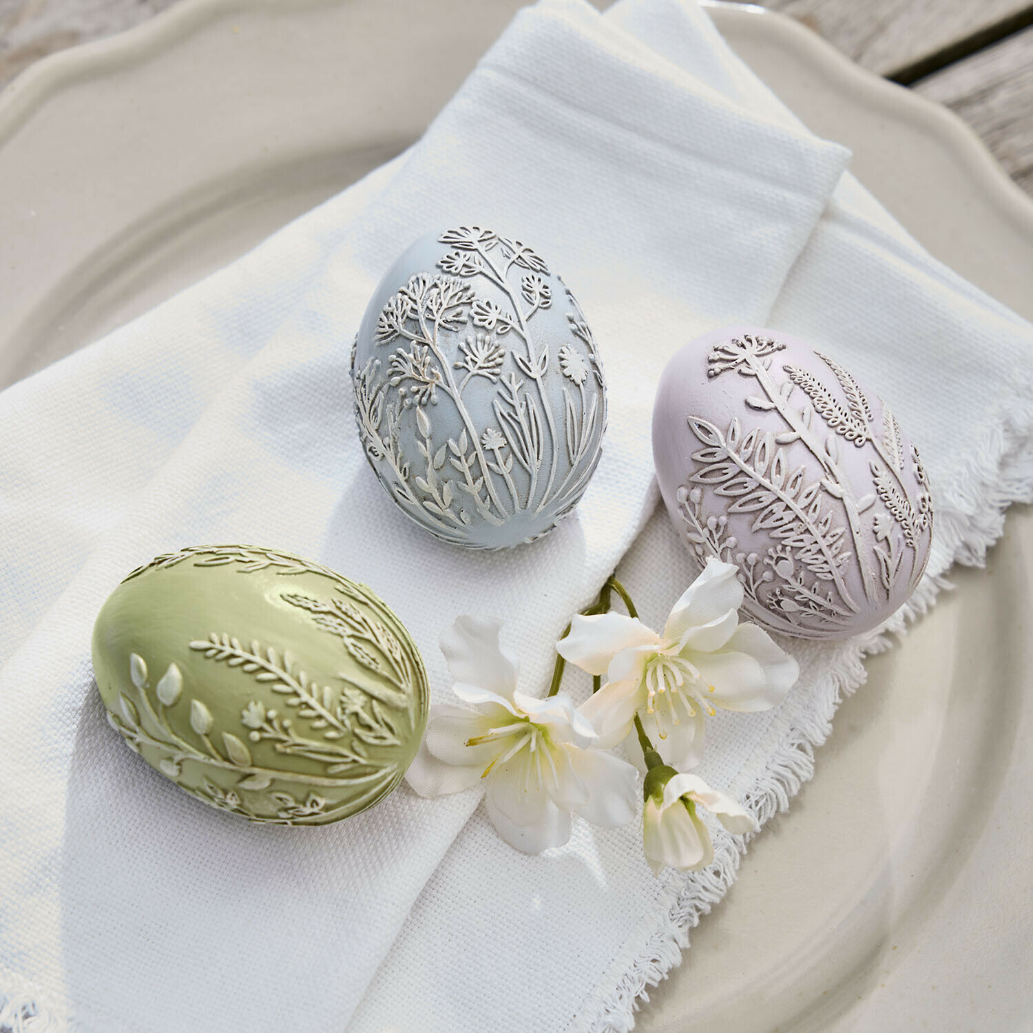 Lot de 3 œufs de Pâques Fauverine Trois œufs décoratifs pastel avec motifs floraux en relief sur tissu blanc et fleurs sur assiette