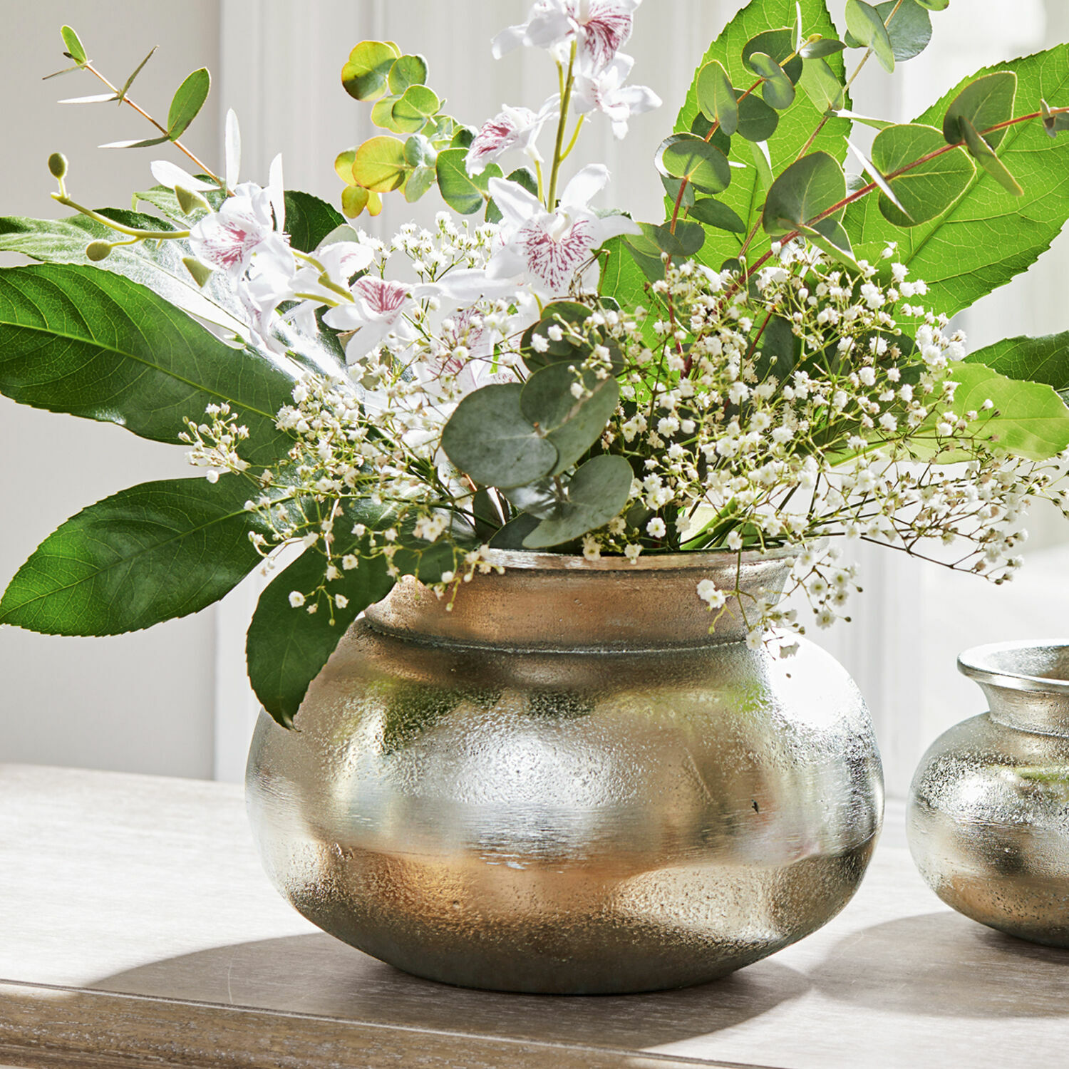 Vase Hadrien Vase en métal argenté avec fleurs blanches et feuillage vert sur table en bois, décoration intérieure