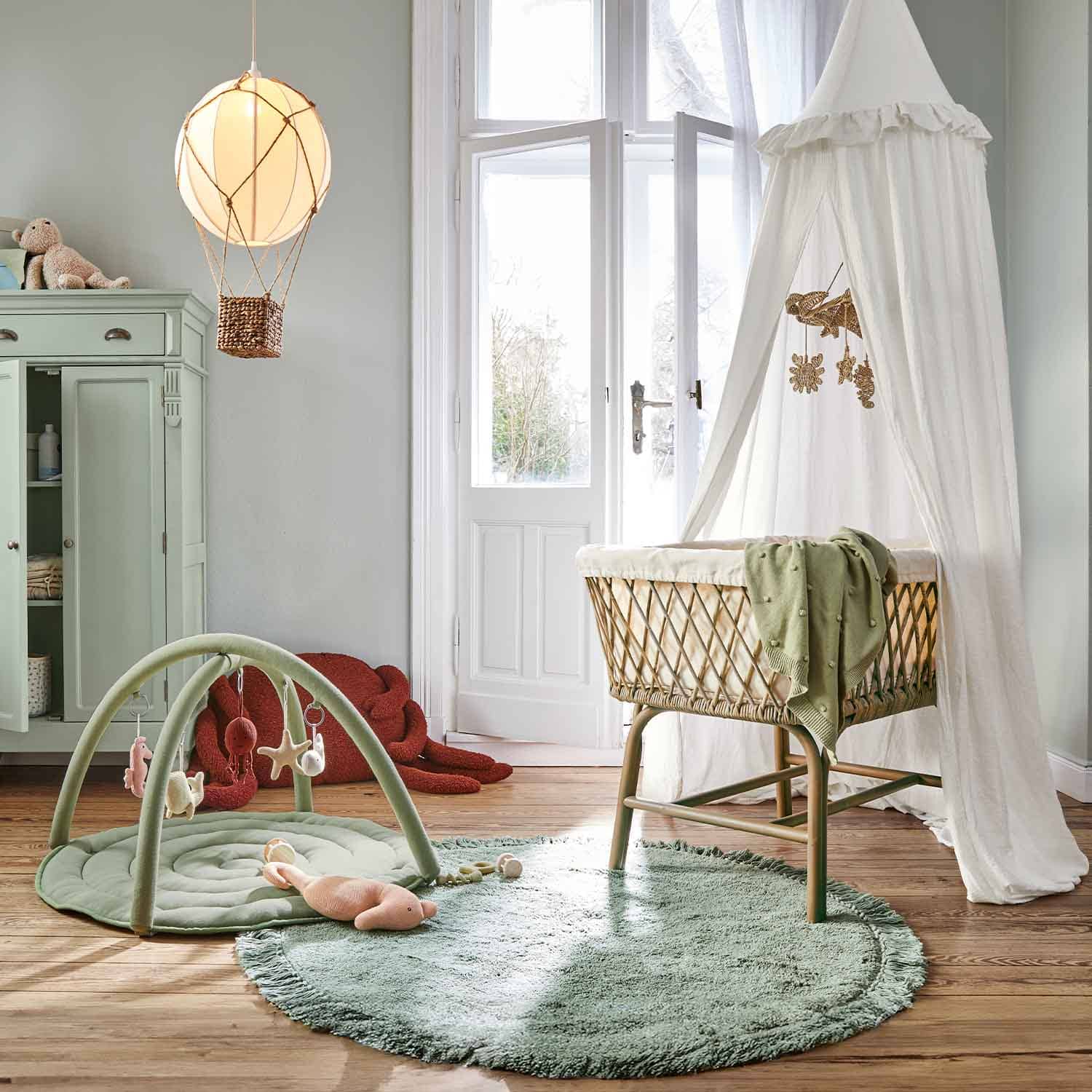 Chambre d'enfant avec berceau en osier et ciel de lit blanc, tapis vert et aire de jeu douce