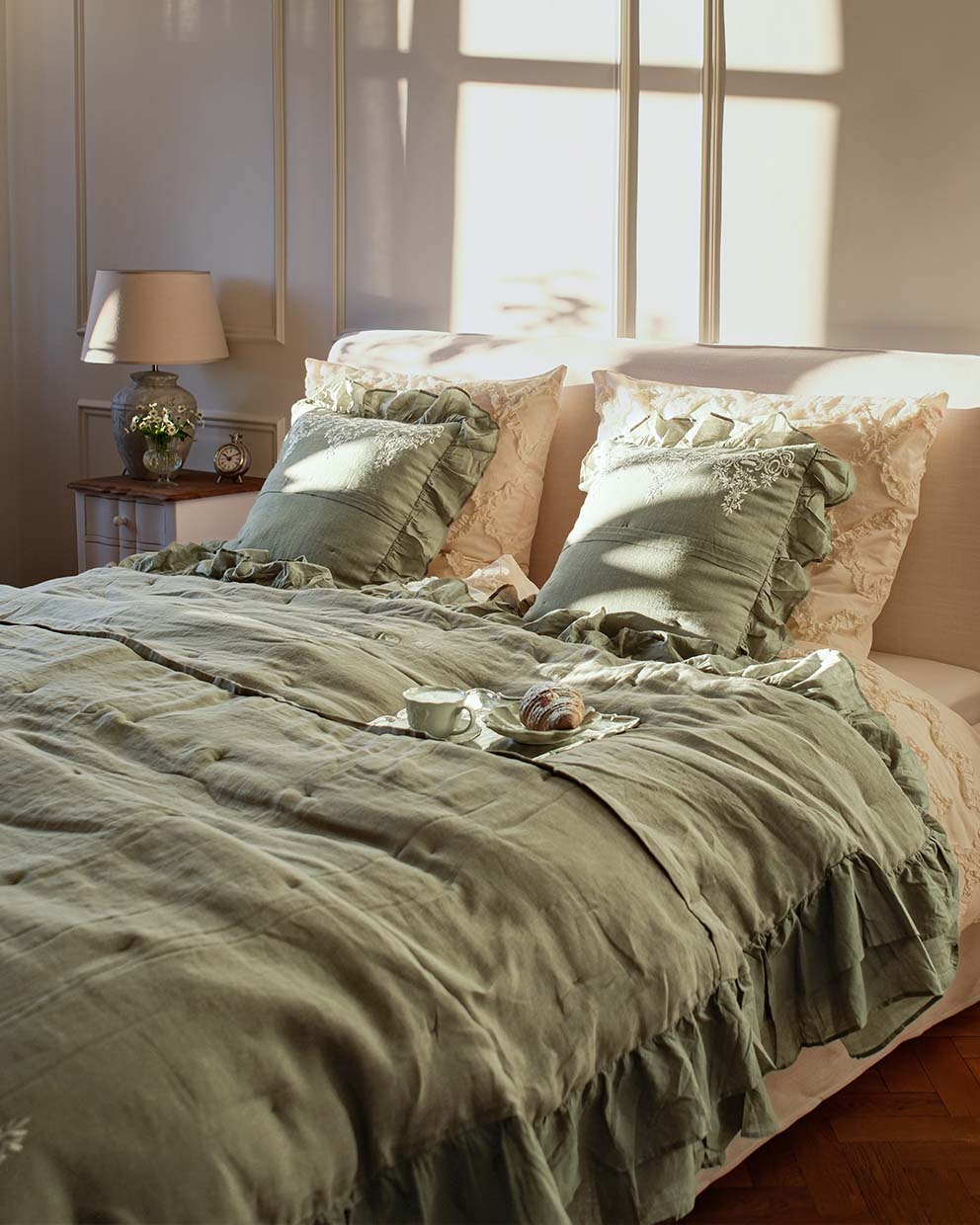 Linge de lit vert en lin avec volants et coussins brodés dans une chambre claire avec lampe et horloge