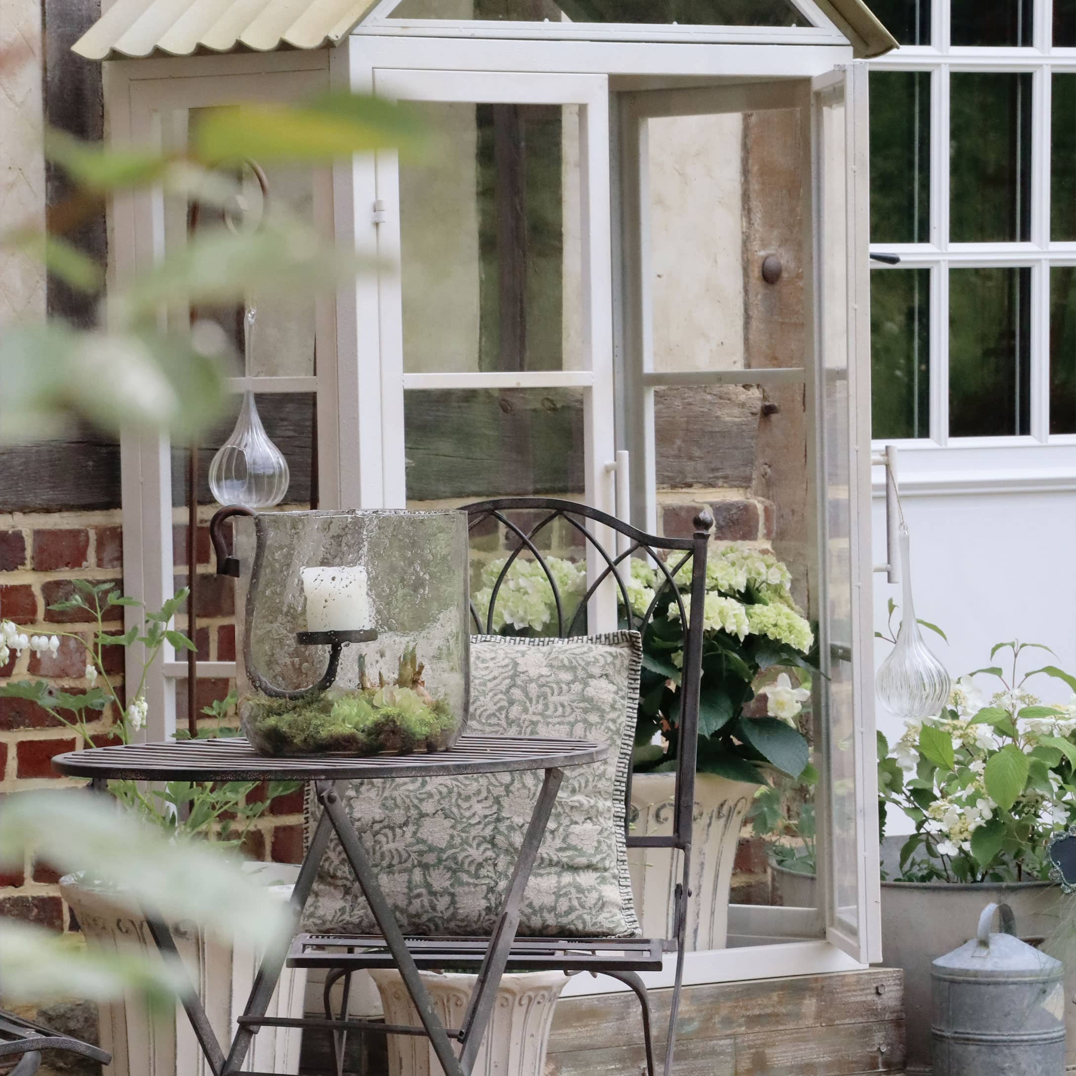 Jardin avec table et chaise en métal, coussin décoratif, lanterne en verre avec bougie blanche et fleurs blanches
