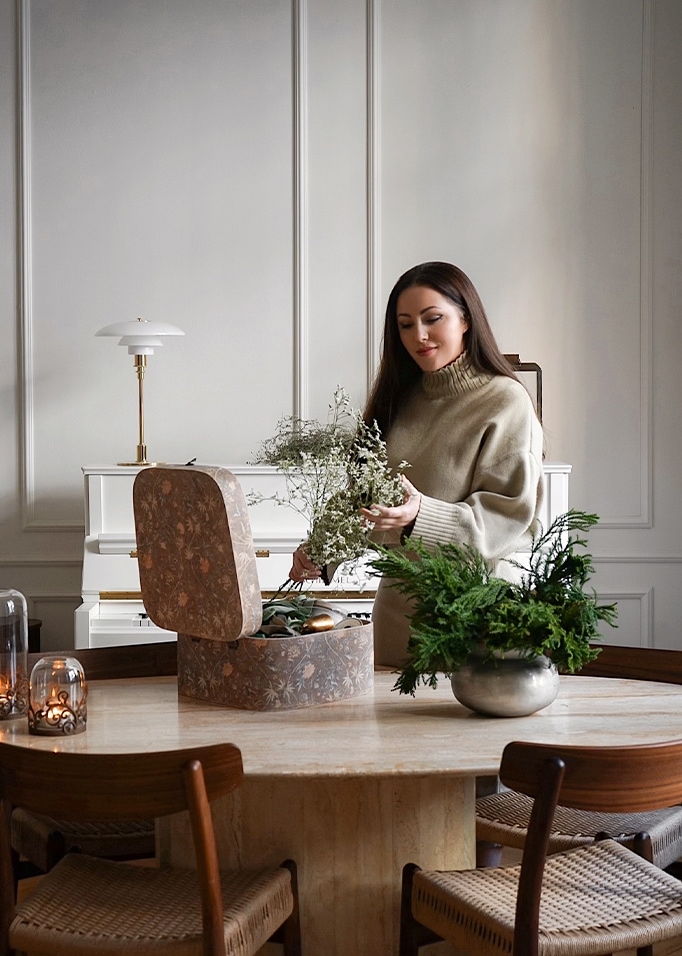 Femme en pull beige arrange des fleurs sur une table en marbre avec boîte décorative et plantes vertes