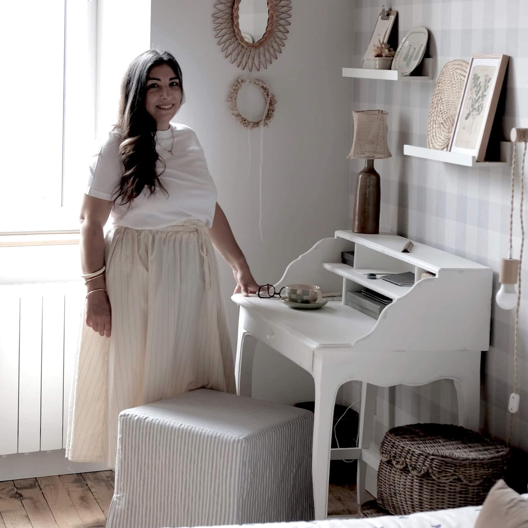 Femme en jupe claire près d'un bureau blanc en bois dans une pièce chaleureuse avec décorations naturelles
