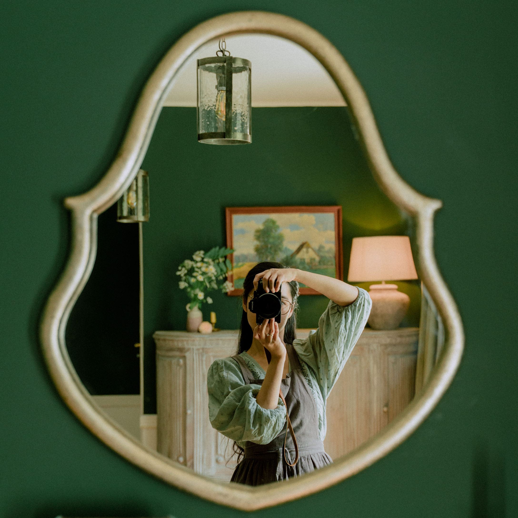 Miroir ovale avec cadre décoratif sur mur vert dans une pièce élégante avec lampe et tableau